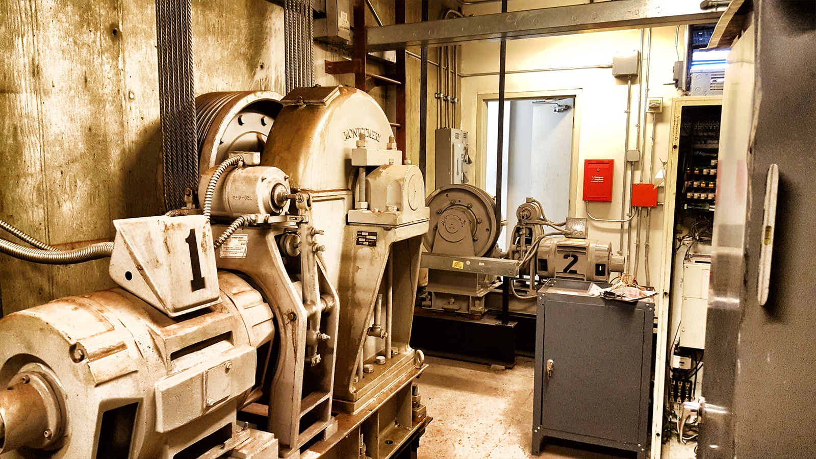 A mechanical room with large industrial elevator machinery, metal equipment, control panels, and electrical boxes amid concrete walls and pipes, lit by overhead lights.